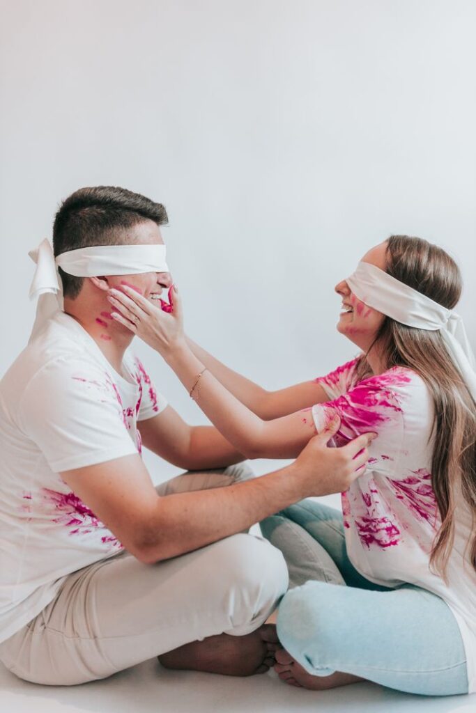 A cheerful couple wearing blindfolds, sitting and smiling, touching each other's faces.